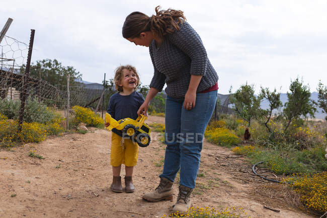 Милый мальчик держит игрушечный бульдозер, стоя с мамой на дорожке на органической ферме. Семья и поместье. — стоковое фото