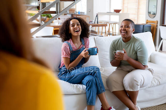 Feliz multirracial amigos do sexo feminino tomando café enquanto sentado no sofá em casa. amizade, socialização e lazer em casa. — Fotografia de Stock