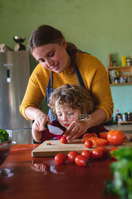 Lindo niño aprendiendo a cortar tomates frescos con cuchillo de la madre en el mostrador de la cocina. alimentación familiar y saludable, estilo de vida doméstico. - foto de stock