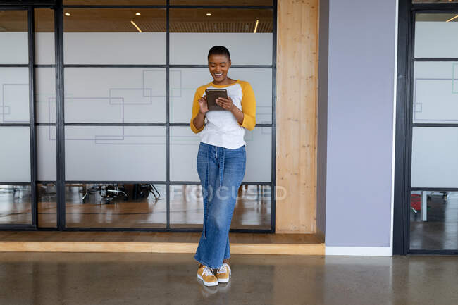Усміхаючись афроамериканська креативна бізнес-леді в нещасних випадках, використовуючи планшетний ПК, стоячи на посаді. креативний бізнес, сучасний офіс та бездротові технології . — стокове фото