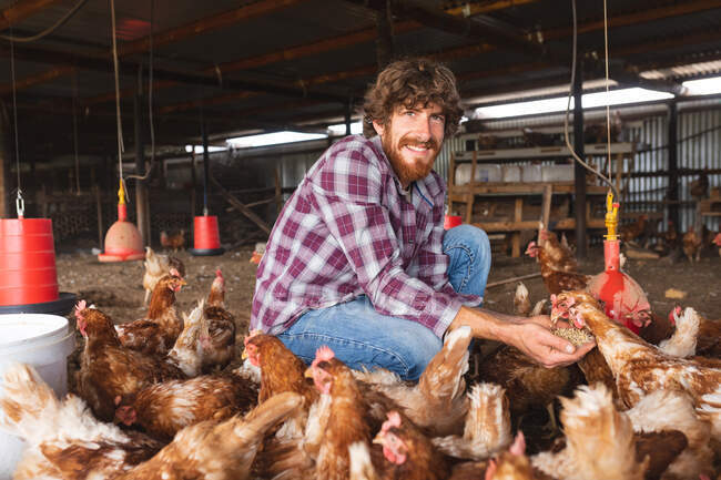 Portrait d'un jeune homme barbu souriant accroupi tout en nourrissant les poules de pastilles en enclos à la ferme. exploitation familiale et avicole, élevage. — Photo de stock