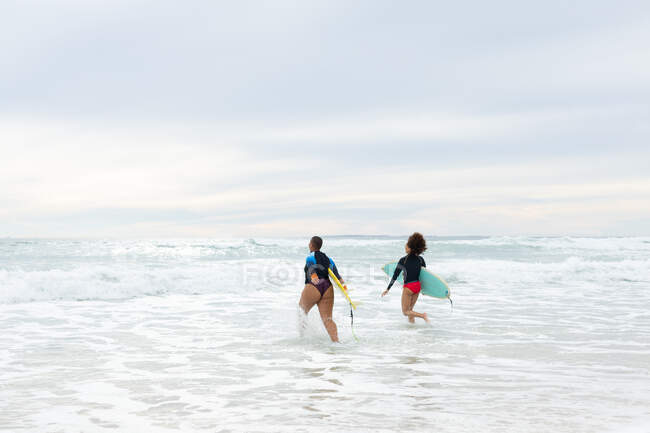 Despreocupadas amigas multirraciales con tablas de surf corriendo en el mar contra el cielo durante el fin de semana. amistad, surf y tiempo libre. - foto de stock