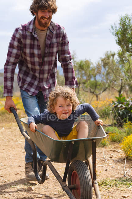 Heureux père barbu poussant son fils assis dans la brouette sur la passerelle à la ferme pendant la journée ensoleillée. famille, homesteading et plaisir. — Photo de stock