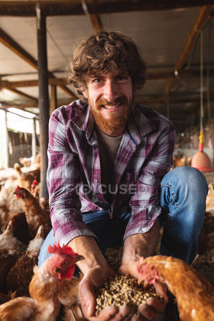 Ritratto di giovane barbuto sorridente che somministra pellet alle galline a penna in fattoria. azienda agricola e avicola, allevamento di bestiame. — Foto stock