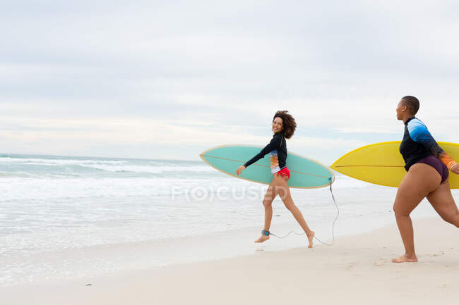 Беззаботные многорасовые подруги с досками для серфинга, работающими на пляже в выходные. дружба, серфинг и досуг. — стоковое фото