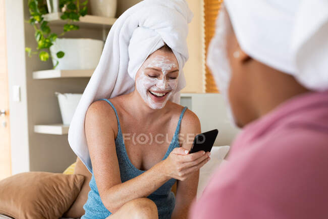 Glückliche Frau mit Gesichtsmaske mit Smartphone, während sie mit Freundin zu Hause sitzt. Freundschaft, drahtlose Technologie und Hautpflege. — Stockfoto