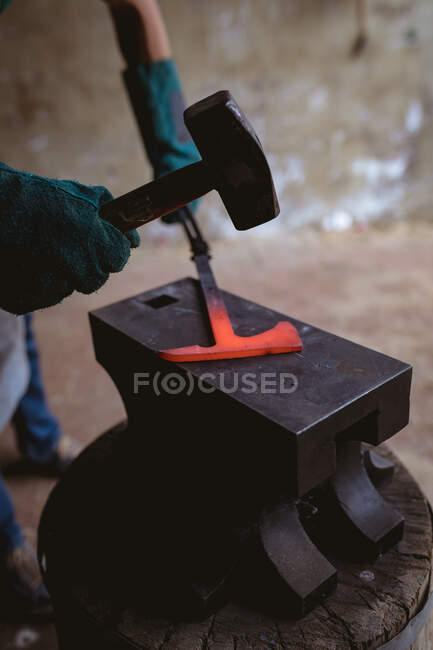 Les mains coupées du forgeron dans les gants de protection forgeant avec le marteau sur l'enclume dans l'industrie. forgeage, métallurgie et industrie manufacturière. — Photo de stock
