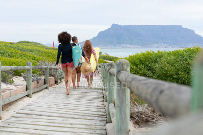 Rear view of female friends carrying surfboards while walking on boardwalk at beach during weekend. friendship, surfing and leisure time. — Stock Photo