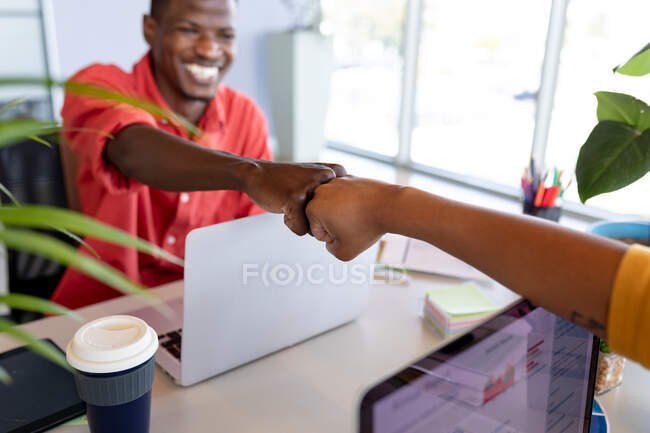 Lächelnder afrikanisch-amerikanischer Geschäftsmann in Kasualien, der einem Kollegen im Kreativbüro einen Faustschlag verpasst. kreatives Geschäft, Büroarbeitsplätze und drahtlose Technologie. — Stockfoto