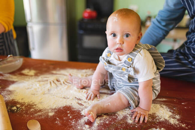 Portrait de bébé mignon jouant tout en étant assis sur la farine sur la table avec les parents dans la cuisine. innocence et famille. — Photo de stock