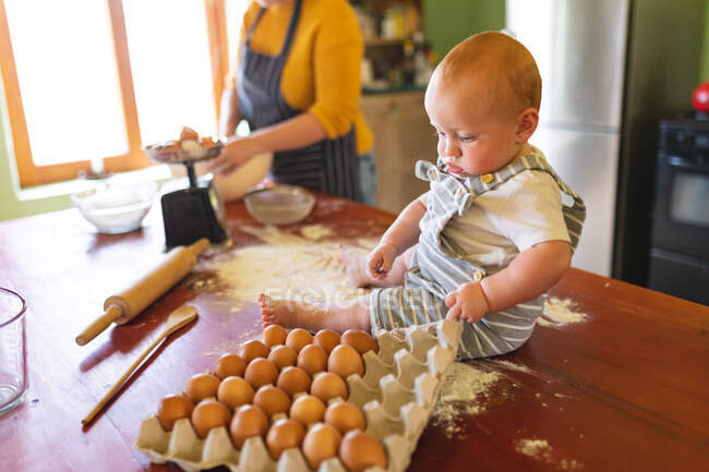 Bebê bonito brincando com a caixa de ovo na mesa de madeira enquanto a mãe prepara comida na cozinha. inocência, família e alimentação saudável. — Fotografia de Stock