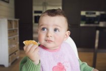 Bebê bonito comer biscoito na cozinha e olhando para longe — Fotografia de Stock