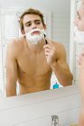 Молодой человек без рубашки бреется перед зеркалом в ванной — стоковое фото