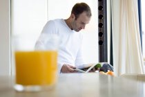 Чоловік читає журнал на кухні, апельсиновий сік на передньому плані — стокове фото