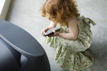 Маленька дівчинка-імбир, прив'язана перед телевізором, тримає пульт дистанційного керування — стокове фото