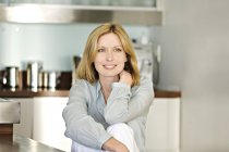 Sonriente mujer pensativa sentada en la cocina y mirando hacia otro lado - foto de stock