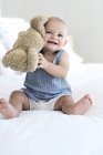 Bebê alegre sentado na cama com ursinho de pelúcia — Fotografia de Stock