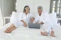 Зріла пара в халатах використовує ноутбук у ліжку — стокове фото
