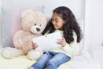 Glückliches kleines Mädchen mit digitalem Tablet und Teddybär im Bett — Stockfoto