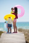 Femme avec ses enfants tenant des anneaux gonflables sur une promenade sur la plage — Photo de stock