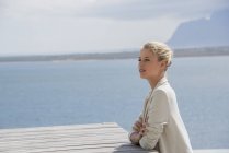 Елегантна молода жінка сидить за столом на березі озера і дивиться геть — стокове фото
