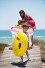 Mujer con sus hijos sosteniendo anillos inflables en un paseo marítimo en la playa - foto de stock