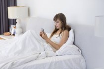 Giovane donna seduta sul letto sotto coperta e utilizzando il telefono cellulare — Foto stock