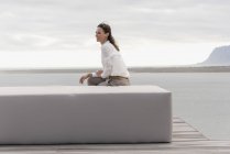 Happy mature woman sitting on ottoman at lake shore and looking away — Stock Photo