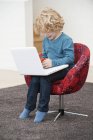 Junge spielt zu Hause im Sessel mit Handy und Laptop — Stockfoto
