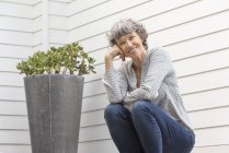 Счастливая зрелая женщина сидит рядом с растением в горшке перед стеной дома — стоковое фото