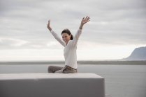 Portrait of happy woman sitting on ottoman with arms raised at lake shore — Stock Photo
