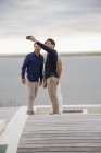 Amigos tomando selfie con teléfono móvil en terraza de madera en el lago - foto de stock