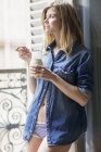 Мрійлива усміхнена жінка в джинсовій сорочці п'є молоко на балконі — стокове фото