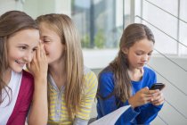 Estudiantes mujeres chismorreando mientras chica solitaria usando el teléfono móvil en la escuela - foto de stock