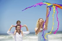 Father carrying little girl on shoulders while mother flying kite on beach — Stock Photo