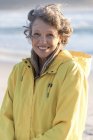 Portrait of happy mature woman in raincoat standing on beach — Stock Photo
