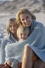 Portrait of a smiling mother and kids wrapped in shawl sitting on beach — Stock Photo