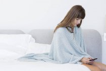 Молодая женщина сидит на кровати и с помощью мобильного телефона — стоковое фото