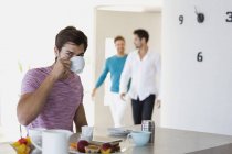 Молодой человек пьет кофе дома с друзьями, идущими на задний план — стоковое фото