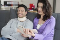 Woman reading book for husband suffering from neck ache — Stock Photo