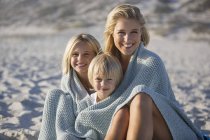Portrait of a smiling mother and kids wrapped in shawl sitting on beach — Stock Photo