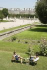 Hombre usando laptop y mujer leyendo una revista en jardín, Jardín des Tuileries, París, Ile-de-France, Francia - foto de stock