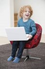 Niño alegre con el pelo rubio usando un ordenador portátil en sillón en casa - foto de stock