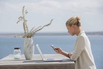Mujer joven usando teléfono inteligente en la mesa de madera con ordenador portátil en la orilla del lago - foto de stock