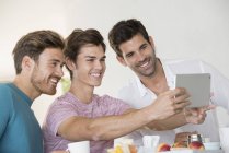 Happy male friends taking selfie with tablet on breakfast table at home — Stock Photo