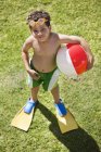 Kleiner Junge in Schwimmflossen und Schwimmbrille hält einen Strandball auf grünem Rasen — Stockfoto
