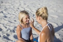 Женщина наносит солнцезащитный крем на нос дочери на песчаном пляже — стоковое фото