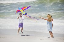 Mutter trägt Kind, während Mann Drachen am Strand fliegen lässt — Stockfoto
