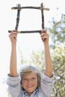Porträt eines niedlichen Jungen mit einem Rahmen aus Treibholz und erhobenen Armen im Freien — Stockfoto