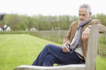 Mensajería de texto de hombre con teléfono móvil mientras está sentado en el banco en el campo - foto de stock